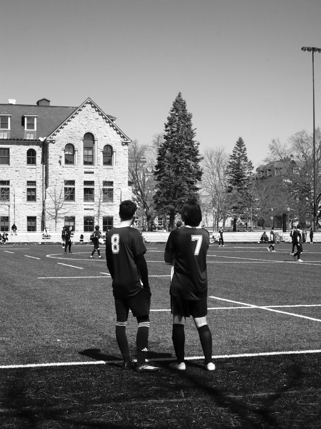 Two soccer players watch a game from an artificial turf field.